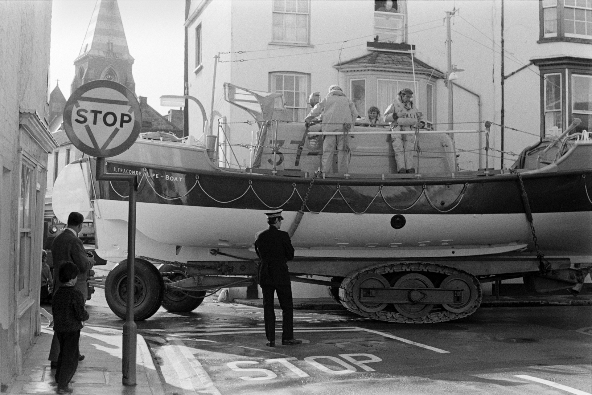 A life boat being transported through Ilfracombe to the harbour on a vehicle with caterpillar tracks. The crew are on the boat and people are watching it from the streets. A policeman is stood at a road junction while the boat moves past.
