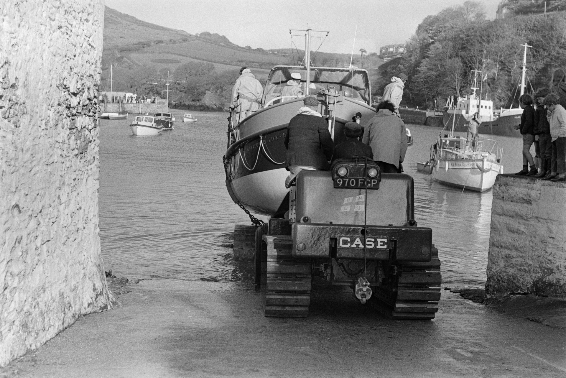 Men launching a life boat into the harbour at Ilfracombe, using a vehicle with caterpillar tracks.  A group of children are watching by the harbour wall.