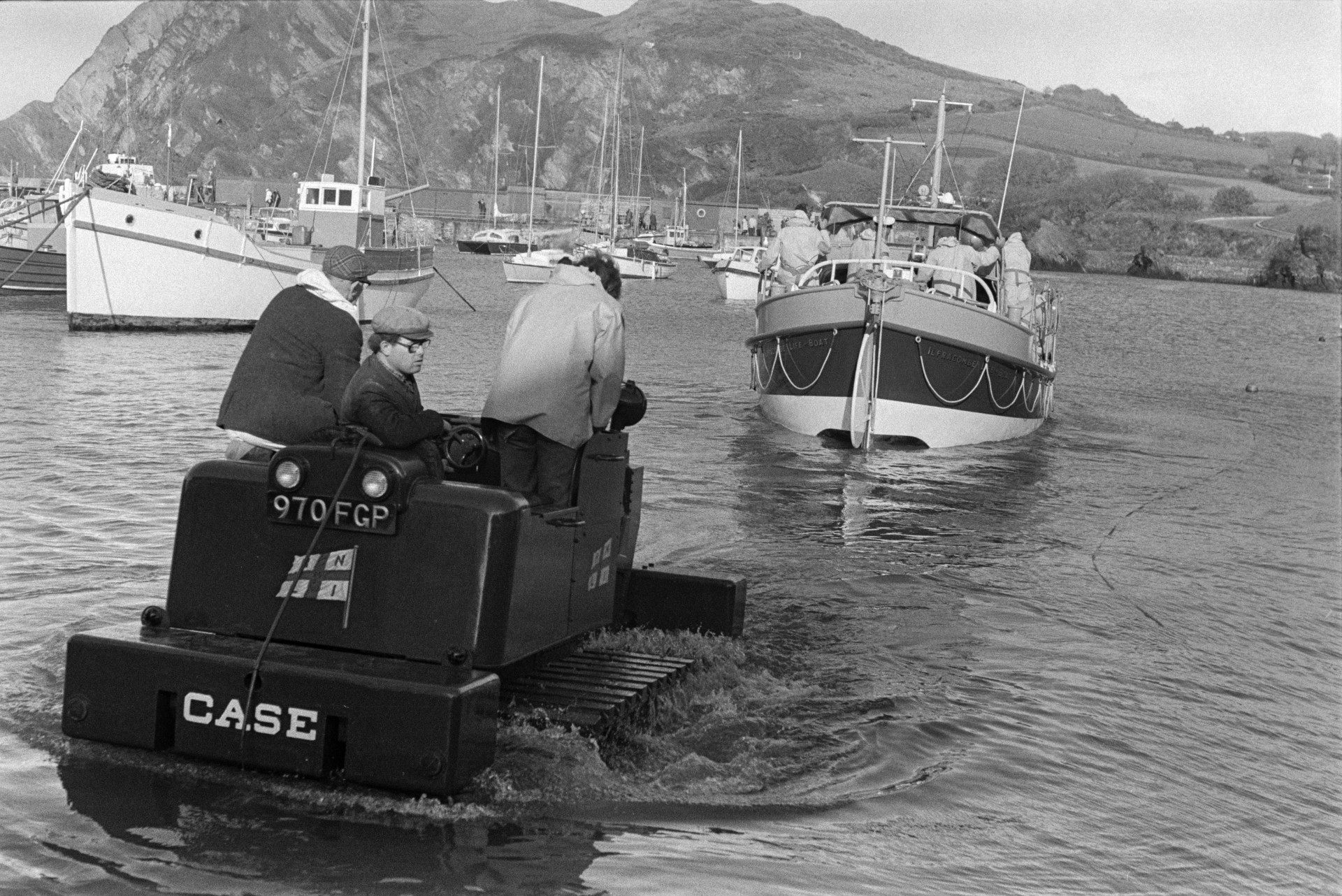 Men launching a life boat into the harbour at Ilfracombe, using a vehicle with caterpillar tracks. Other boats can be seen moored in the harbour.