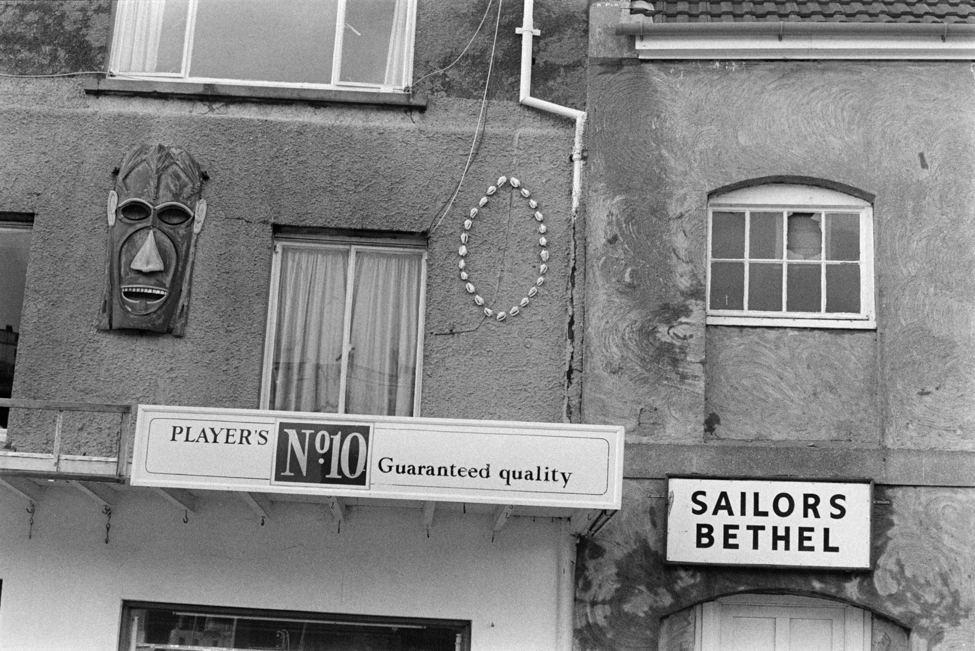 The exterior of the Sailor's Bethel or chapel at Ilfracombe. A shop next door has a sign advertising Players No.10 cigarettes, a pattern of shells and a carved wooden mask.