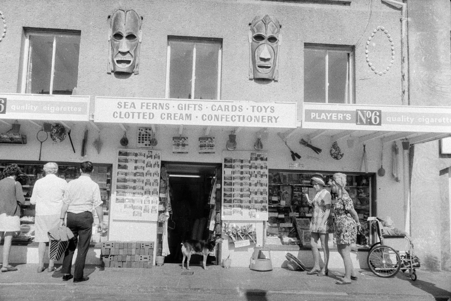 Men and women looking at a gift shop on the seafront at Ilfracombe. Postcards are on display, a dog is stood in the doorway and a woman is sat in a wheelchair outside. An advert for Players No.10 cigarettes is above the shopfront; and a shell pattern and carved wooden masks decorate the shop front.