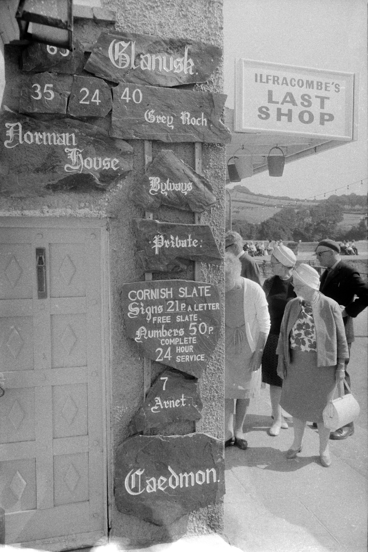 Men and women looking in the shop window of 'Ilfracombe's last shop'. House signs and numbers are displayed outside.