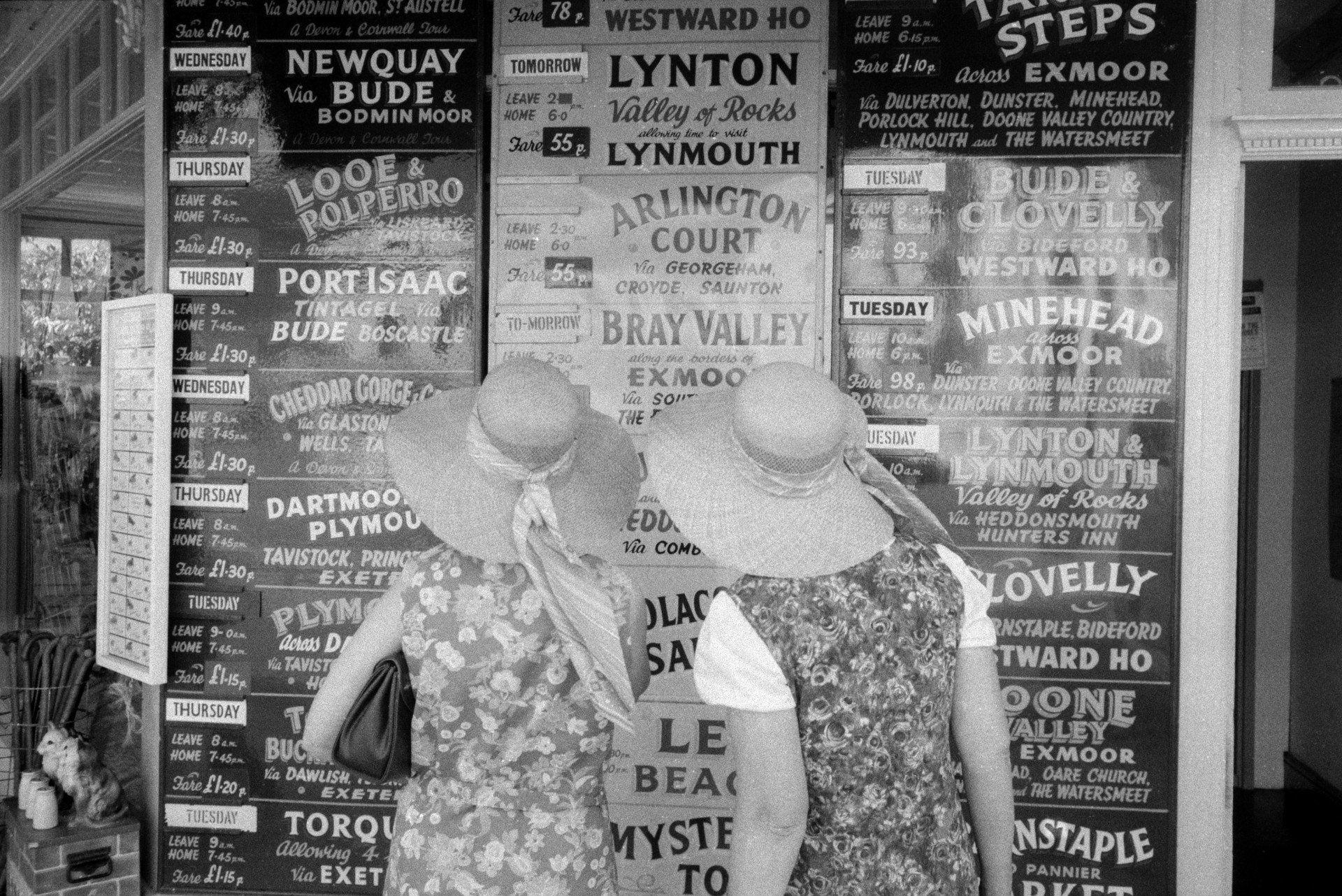Two women wearing summer dresses and hats looking at the adverts for boat trips at Ilfracombe.