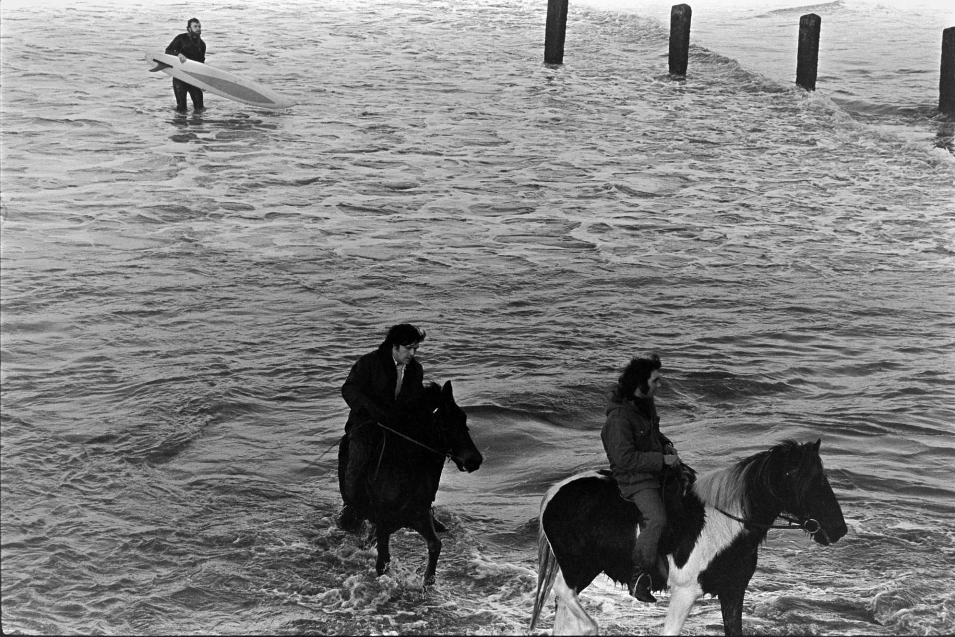 Two men riding horses along the shoreline in the sea, by a sea groyne, at Westward Ho! A surfer is following them out of the sea.