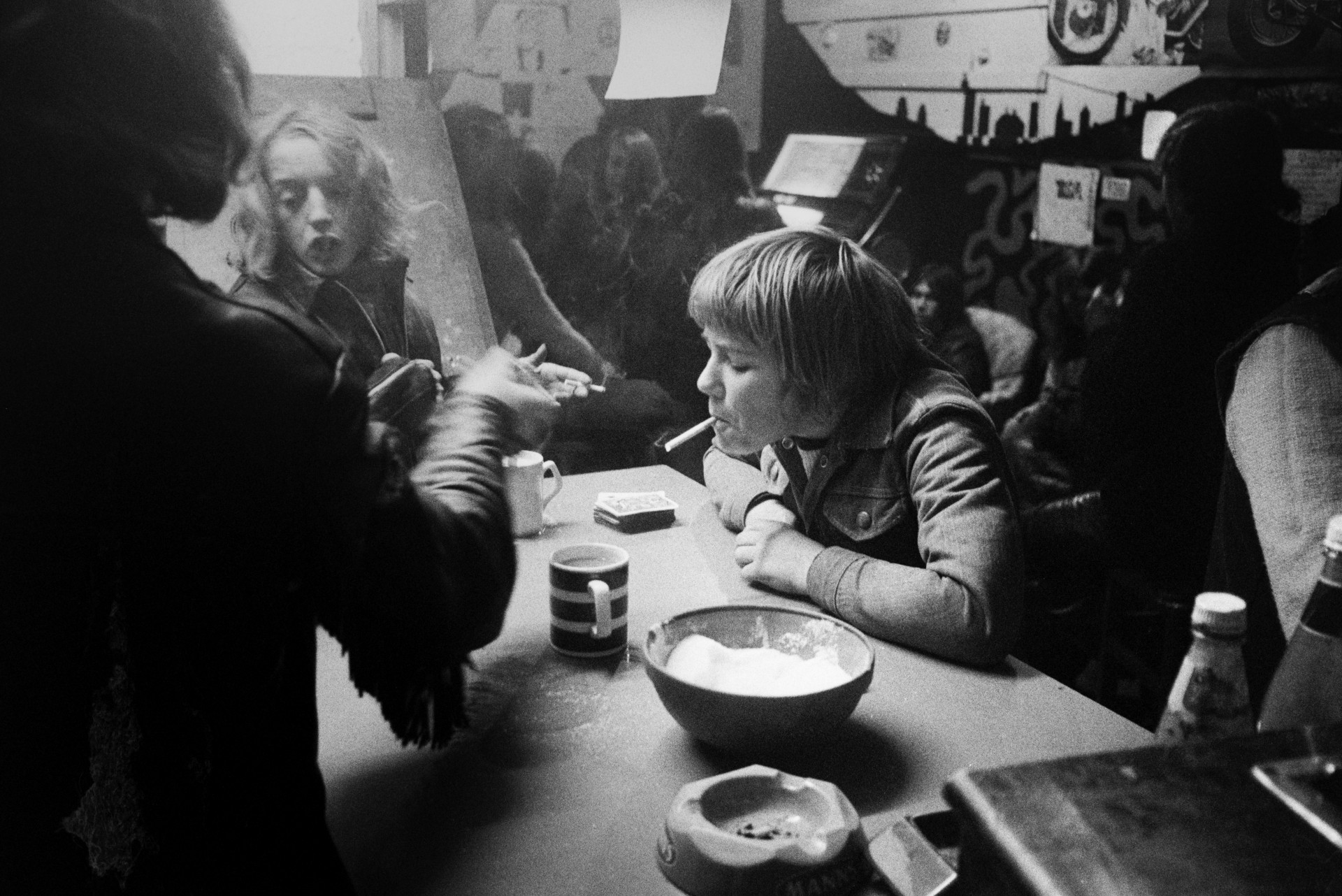 Young men and ta boy playing cards and smoking at a table, possibly in a pub, youth club or café. Mugs and an ashtray are on the table.