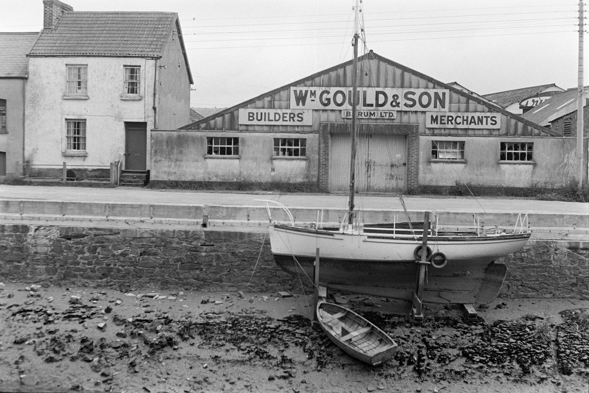 William Gould & Son Builders Merchants premises on the banks of the River Taw at Barnstaple. A moored boat and rowing boat are on the mud flats of the river.