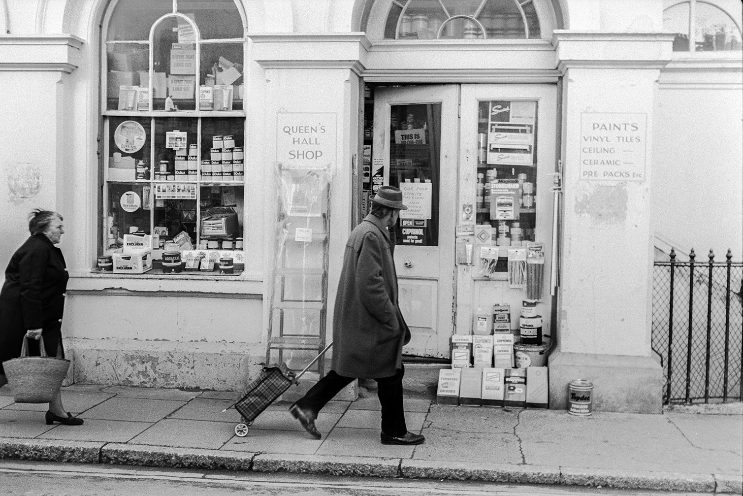 A man and woman walking past the 'Queens Hall Shop' in Barnstaple. Various goods, including tins of paint, are on display outside the shop and in the shop window.
