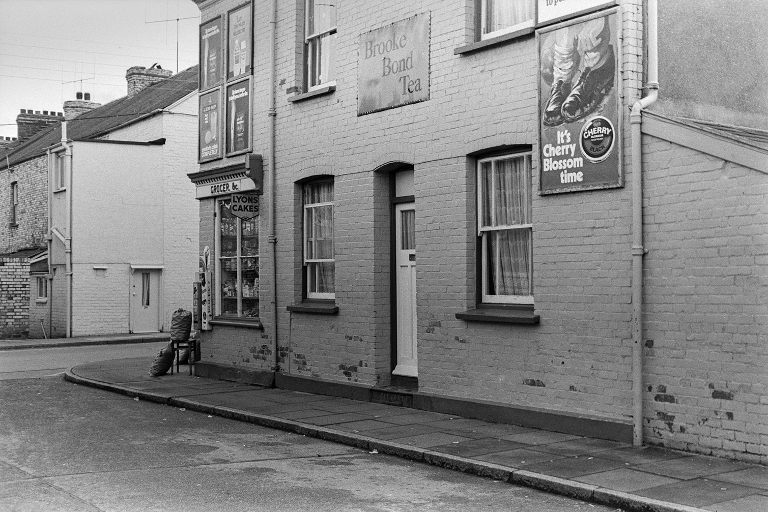 Advertising boards fixed to a Grocer's shop and a house on a street corner in Barnstaple. Adverts include one for cherry blossom shoe polish and Brooke Bond Tea.