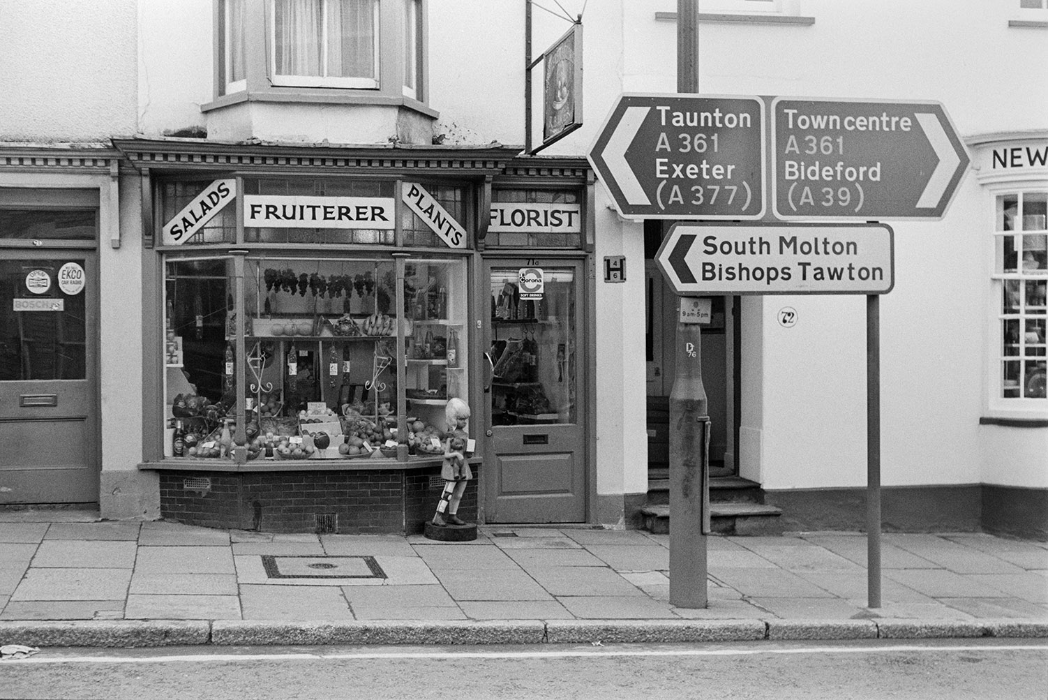 The window of a florist and fruit shop in Barnstaple, with a road sign outside. Apples, bananas and melons are on display in the window.