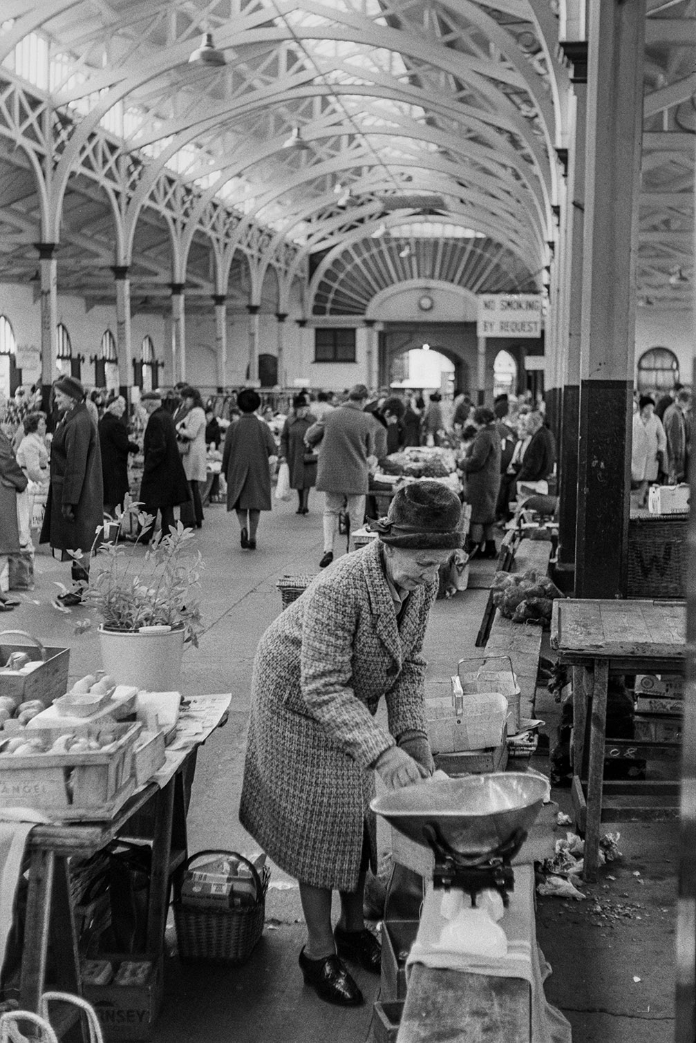 A woman setting out items for sale on a stall at Barnstaple Pannier Market. A set of scales is next to her and eggs and foliage can be seen on the stall. Other stallholder and shoppers can be seen in the background.
