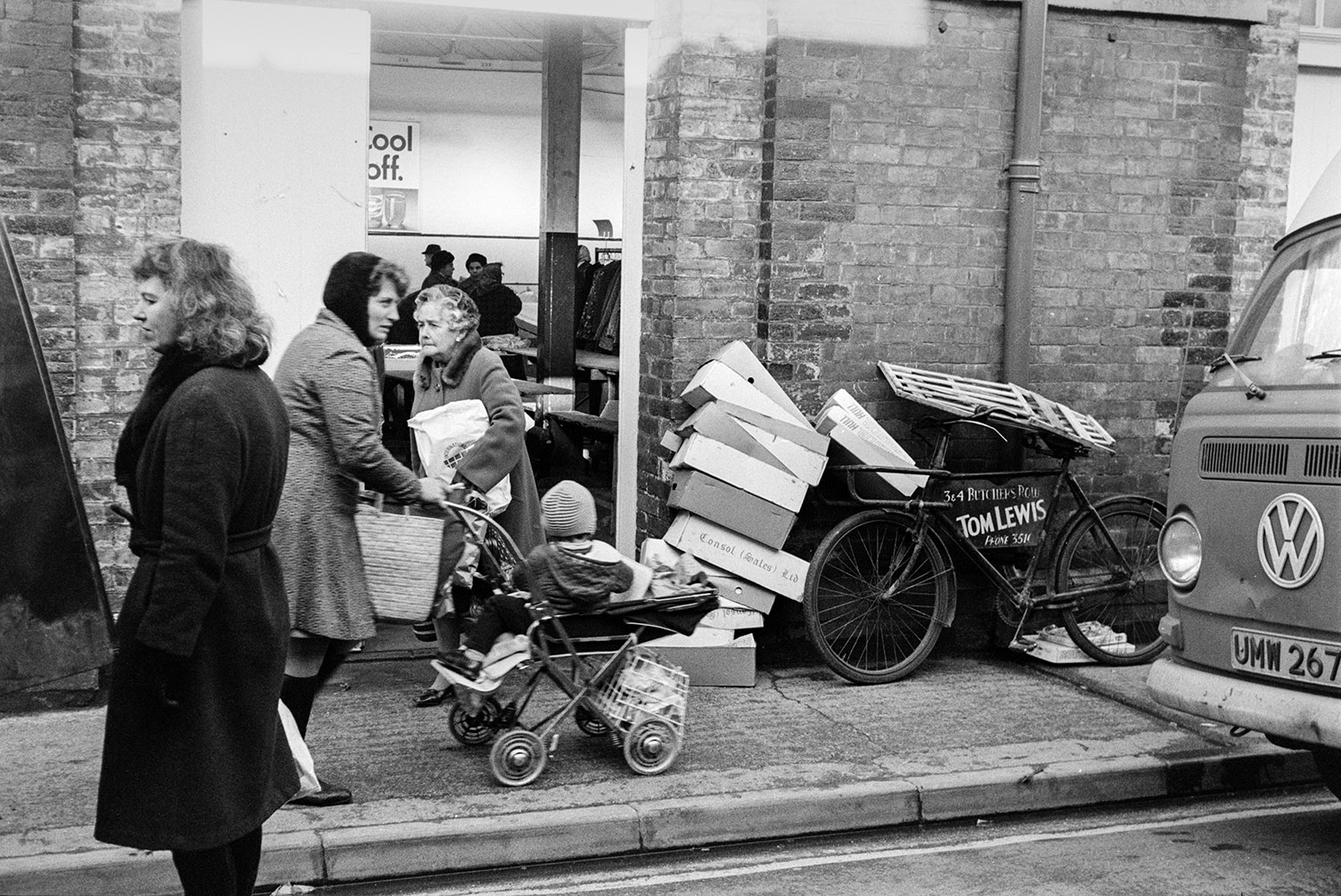 Women walking along a street outside Barnstaple Pannier Market. One woman is pushing a pram with a child, past some cardboard boxes and a parked bicycle.
