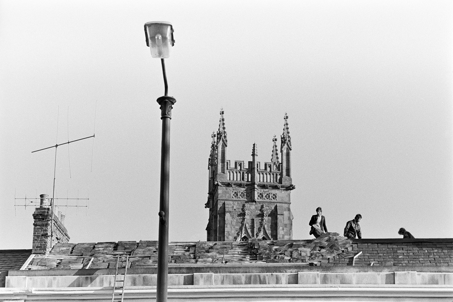 Three men working on the slate roof of terraced houses in Barnstaple. A church tower is visible in the background and  street lamp is in the foreground.