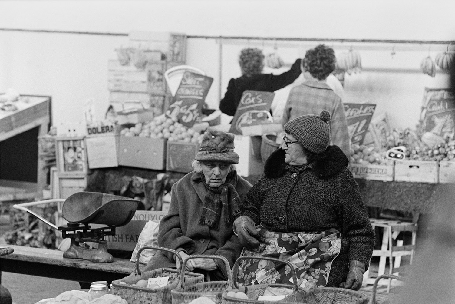 Two women sat down talking and running a stall at Barnstaple Pannier Market. Their good are in baskets in front of them. A stall selling fruit is in the background.