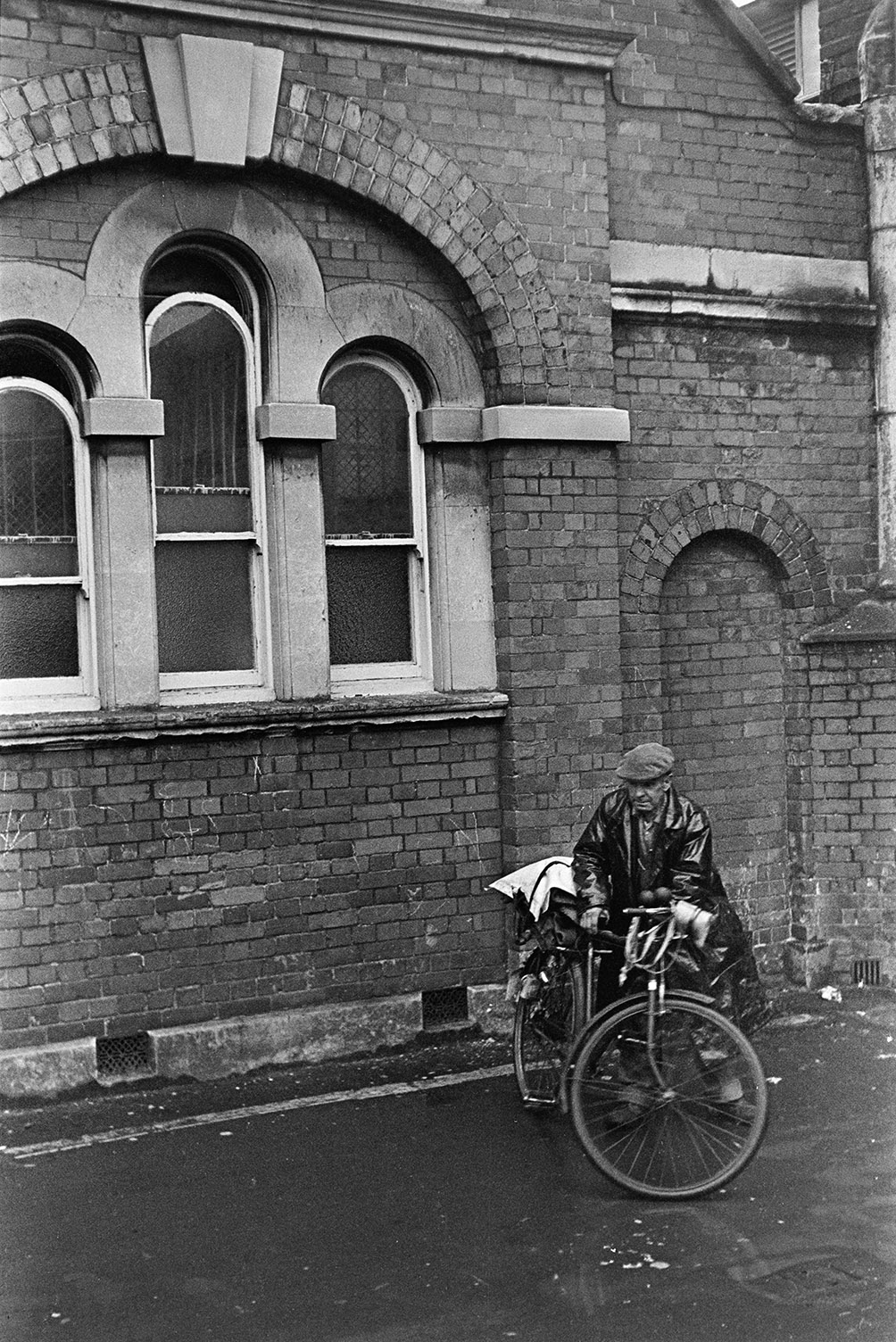 A man with a bicycle outside a building with arched windows in Barnstaple. He is wearing a raincoat.