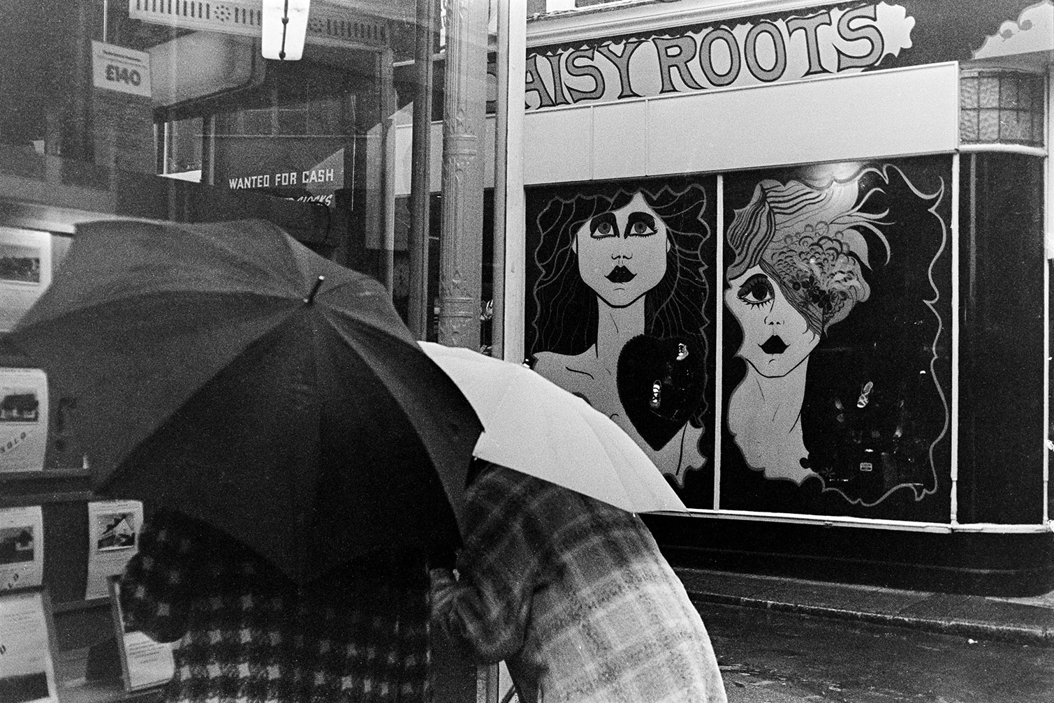 Two people with umbrellas looking in a estate agent's shop window in Barnstaple. Opposite is the shop window of Daisy Roots show shop, with artwork.