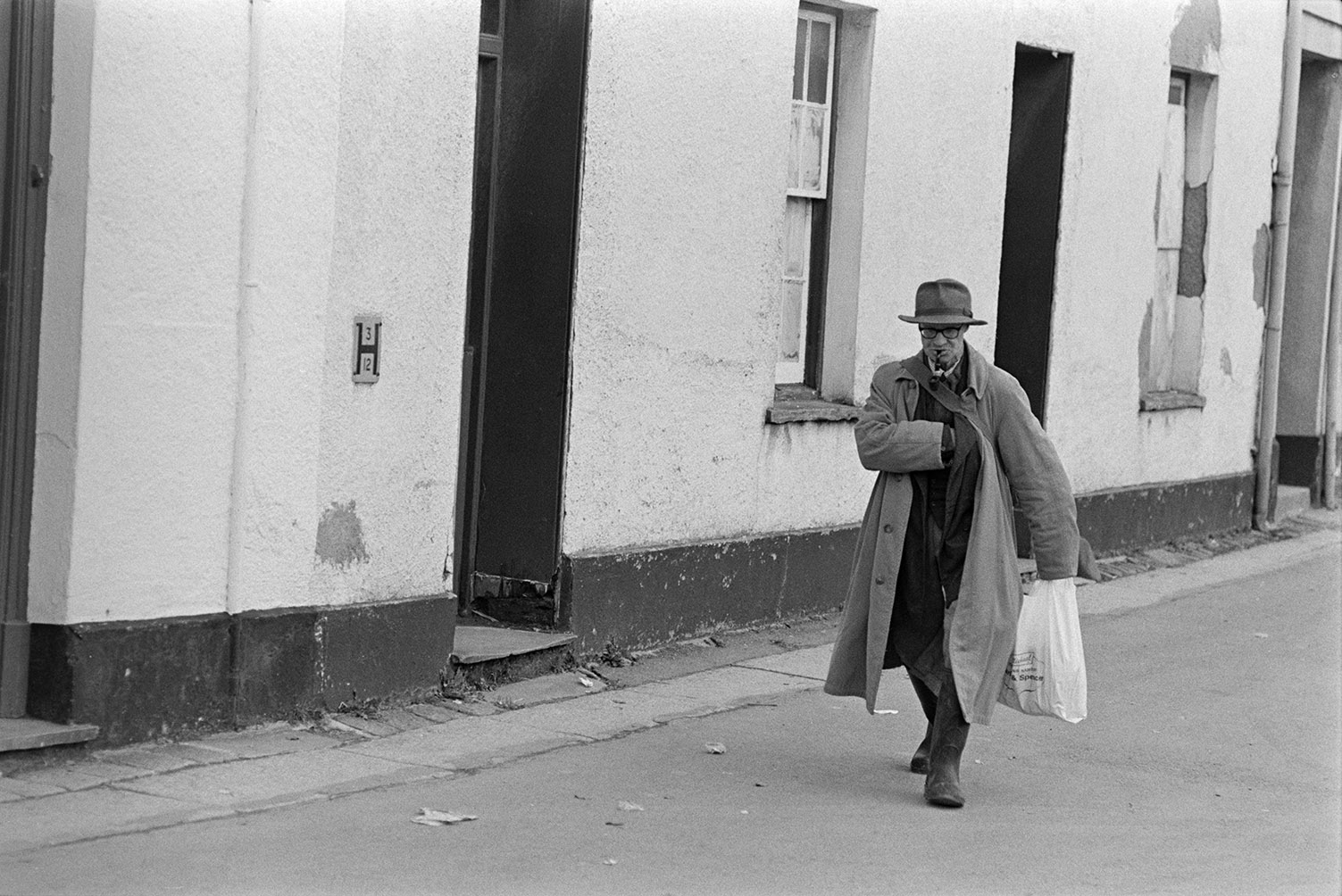 A man walking down a street in Barnstaple. He is smoking a pipe and holding a carrier bag.