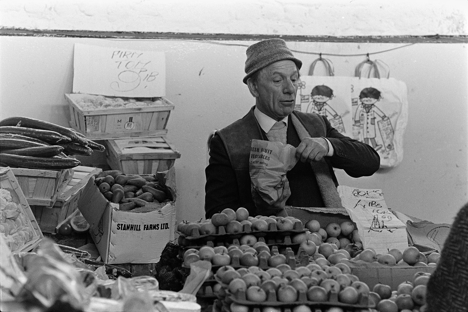 A man running a fruit and vegetable stall at Barnstaple Pannier Market. He is bagging up apples. Cucumbers and pears can also be seen on the stall.