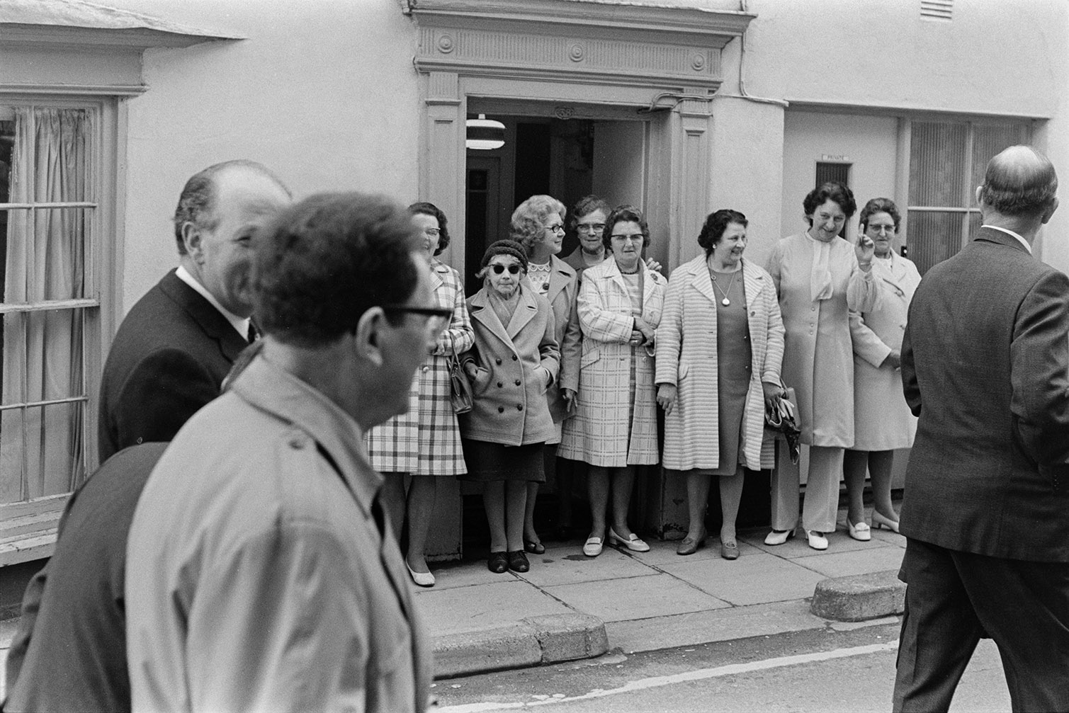 Women gathered by a doorway watching dignitaries walk past in the Torrington May Fair parade.