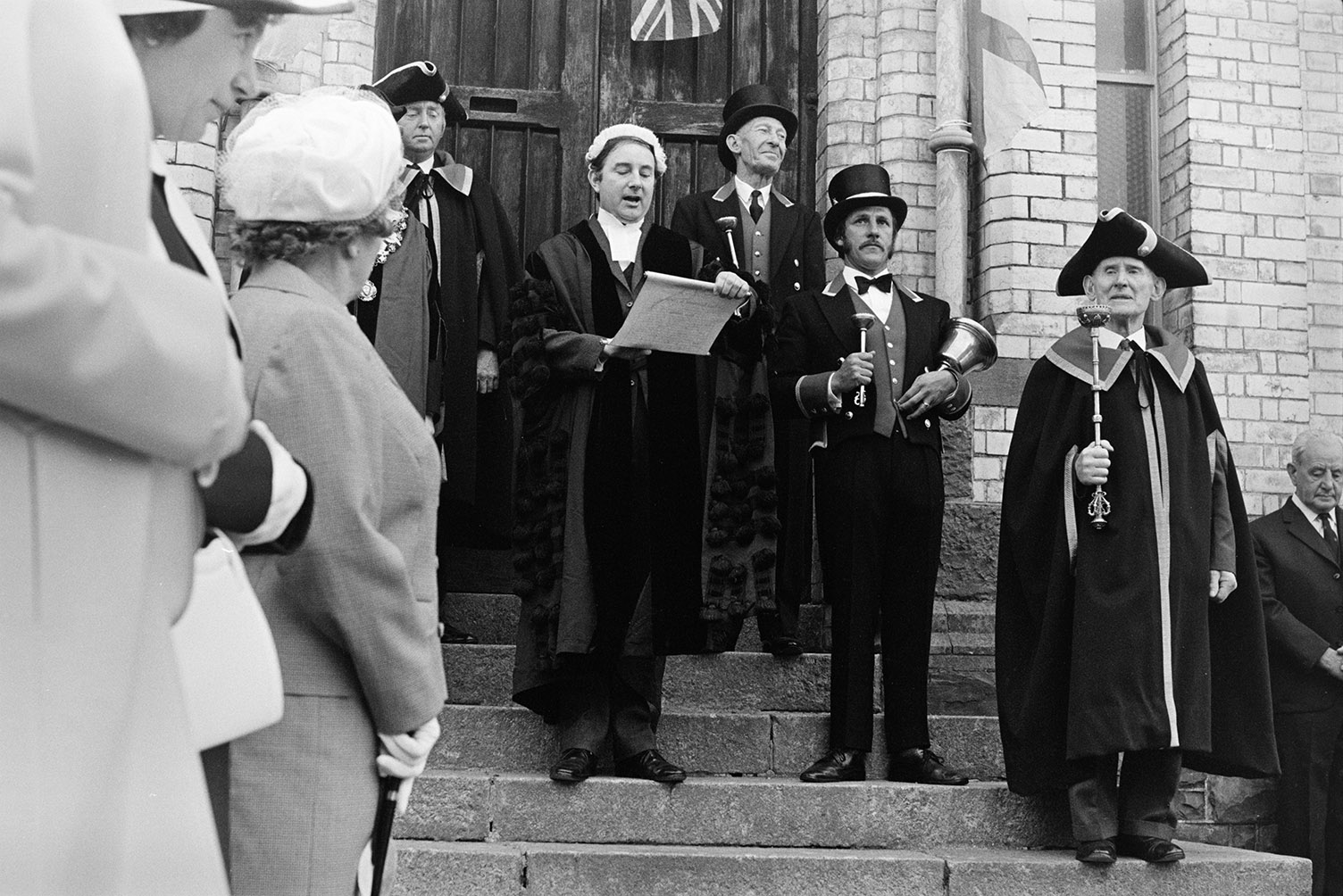 A dignitary giving a speech to a crowd at the Torrington May Fair, from the steps of Vaughan Trapscott Glove Factory in Torrington. Various other dignitaries are listening, including a man holding a mace.