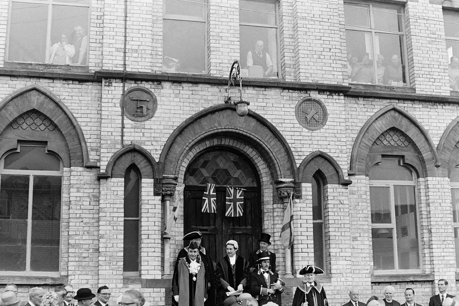 Dignitaries assembling on the steps of Vaughan Trapscott Glove Factory at Torrington May Fair before giving a speech. Union Jack flags are hung above them and people are watching from the first floor windows of the factory.