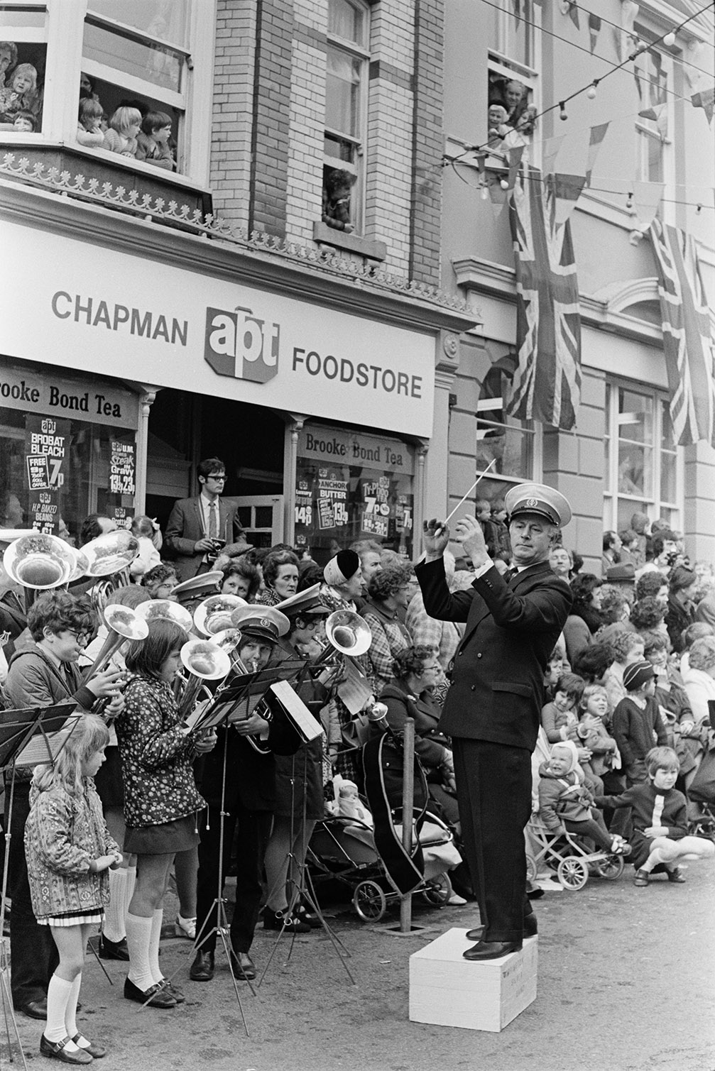 A man conducting a children's brass band at Torrington May Fair outside Chapman Food Store. Spectators are watching the parade from the street and first floor windows of buildings along the street.