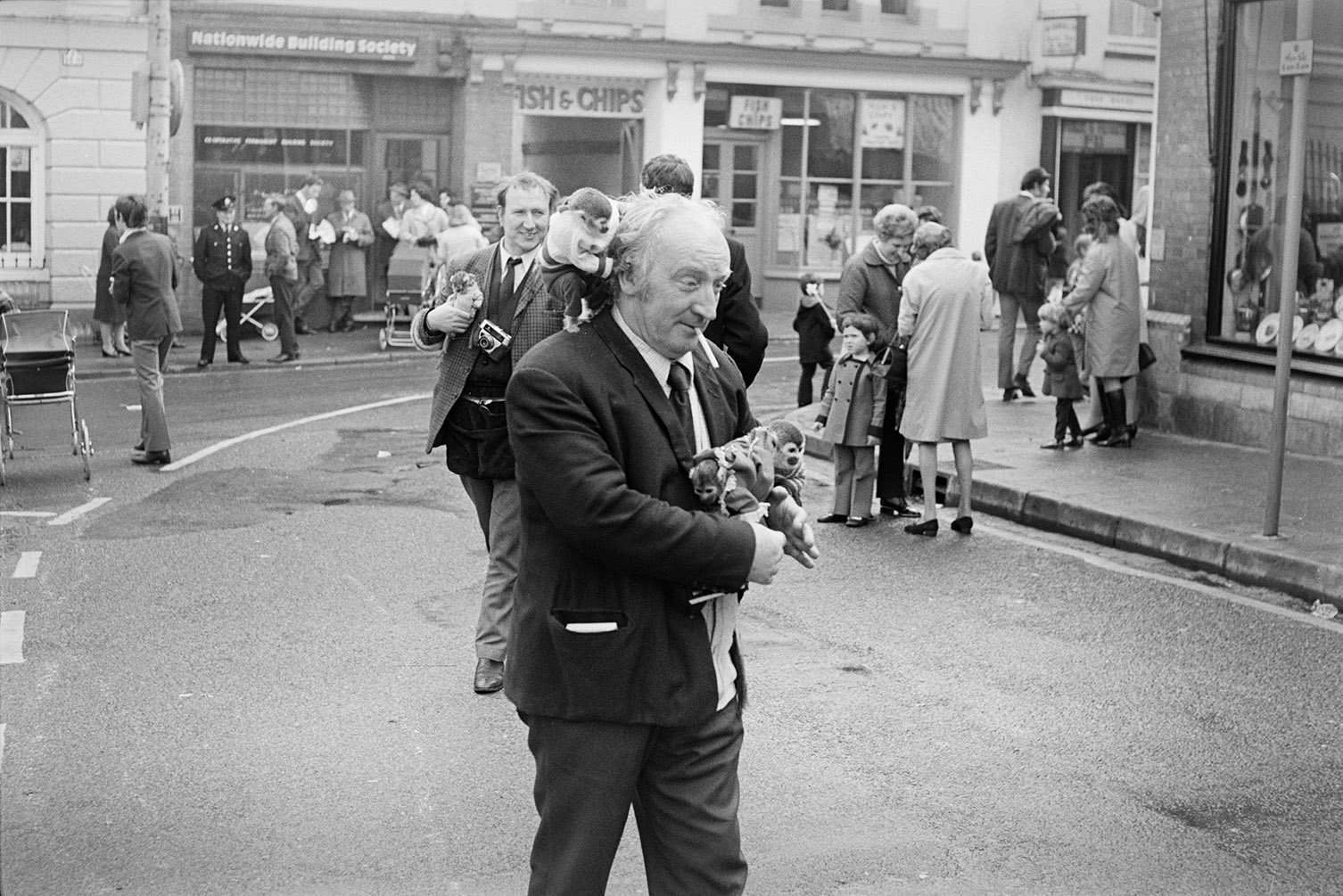 Two men carrying monkeys at Torrington May Fair. One of them is smoking a cigarette and the other has a camera. The Nationwide Building Society and Fish & Chip shop can be seen in the background.