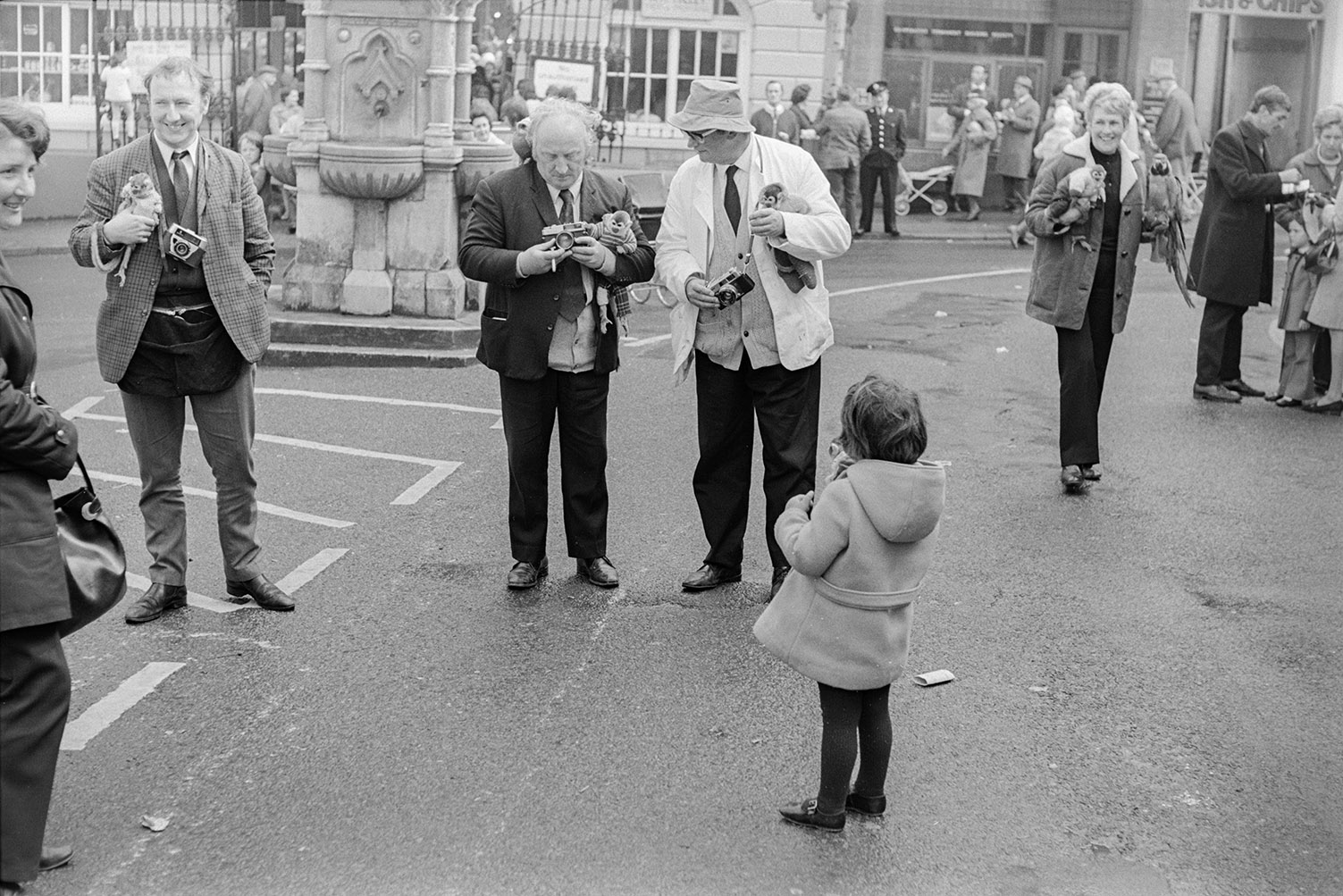 Three men with monkeys and camera at Torrington May Fair. They are taking a photograph of a girl holding a monkey. A woman in the background is carrying a monkey and a parrot.