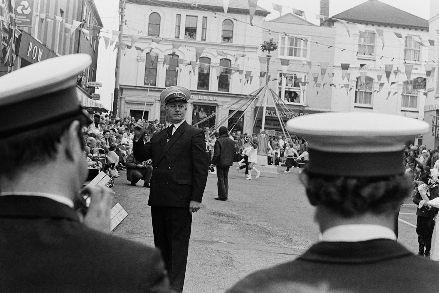 A man conducting a children's brass band at Torrington May Fair. May pole dancers are in the square in the background. The street is decorated with bunting.