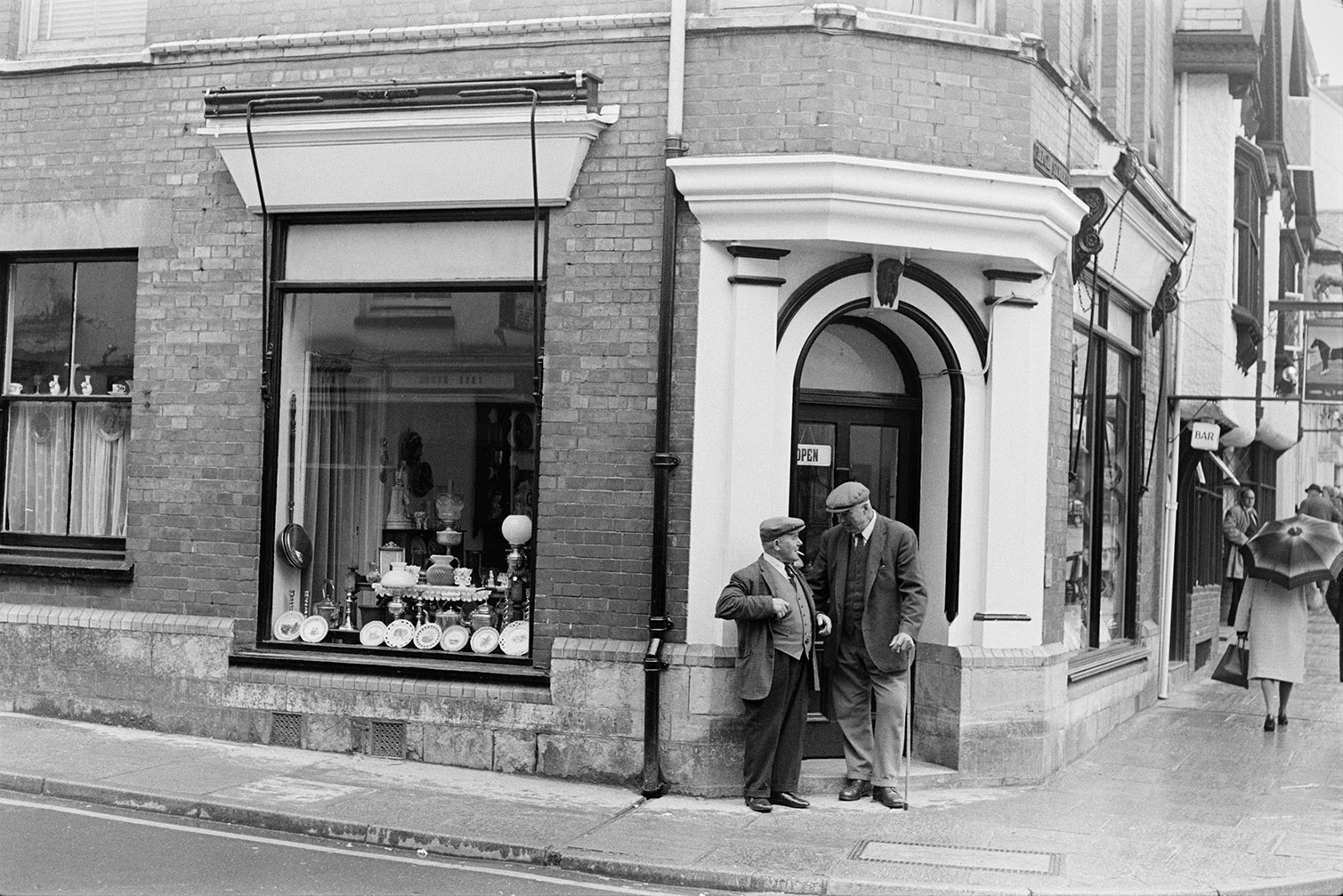 Two men talking outside a shop front window on a street corner in Torrington. The shop is selling antiques including plates, oil lamps and a bed warmer.