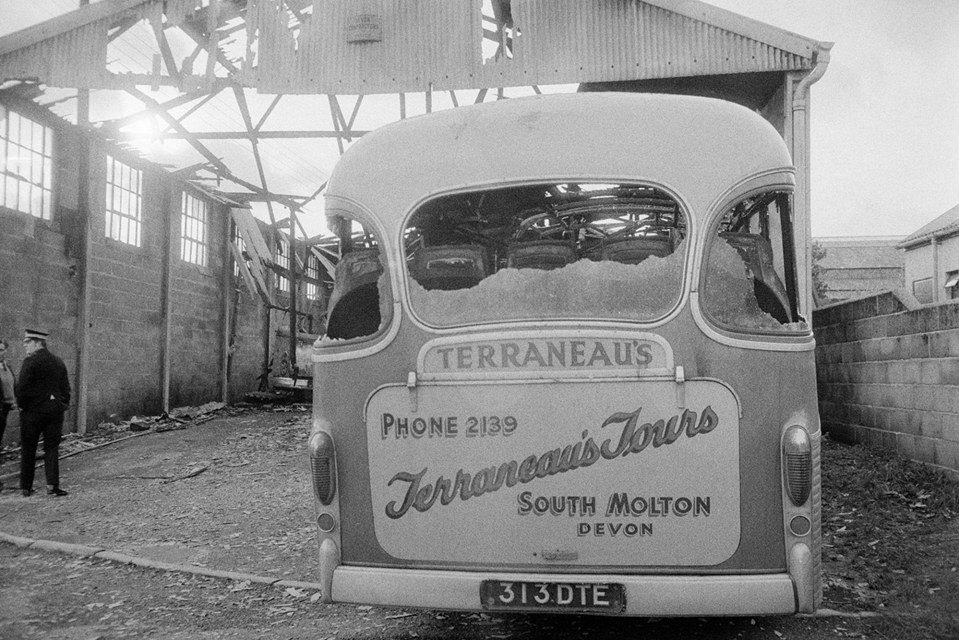 A Terraneau's Tours bus with smashed windows in a burnt out bus depot at South Molton. A policeman and man are talking in the foreground.