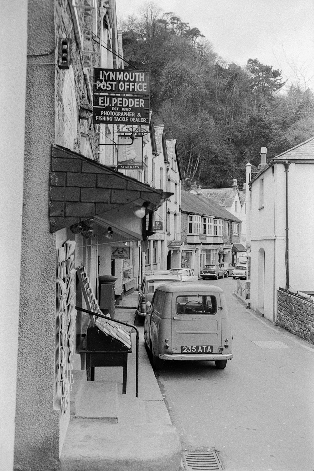View of a street with parked cars and Lynmouth Post Office. A sign on the post office building reads 'E J Pedder photographer & Fishing Tackle Dealer'. Newspapers are displayed on racks outside the Post Office.