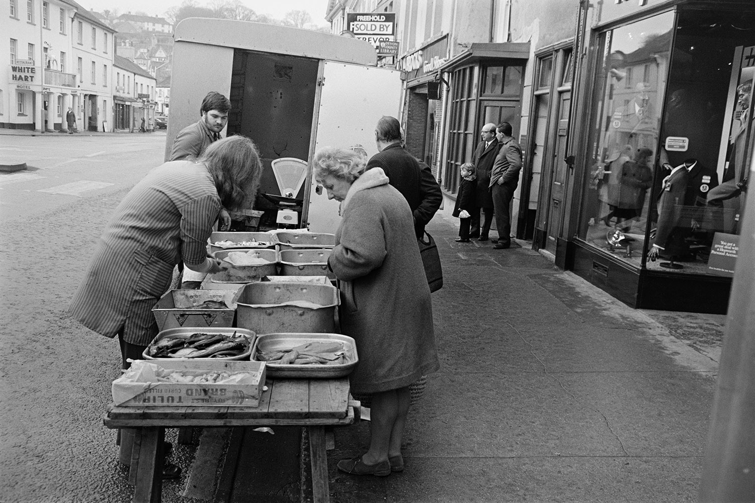Market stalls in a street in Okehampton. A woman is buying from one of the traders who is running a fish stall. Two men and a child are stood outside a shop front in the background and the White Hart Hotel is visible on the opposite side of the street.