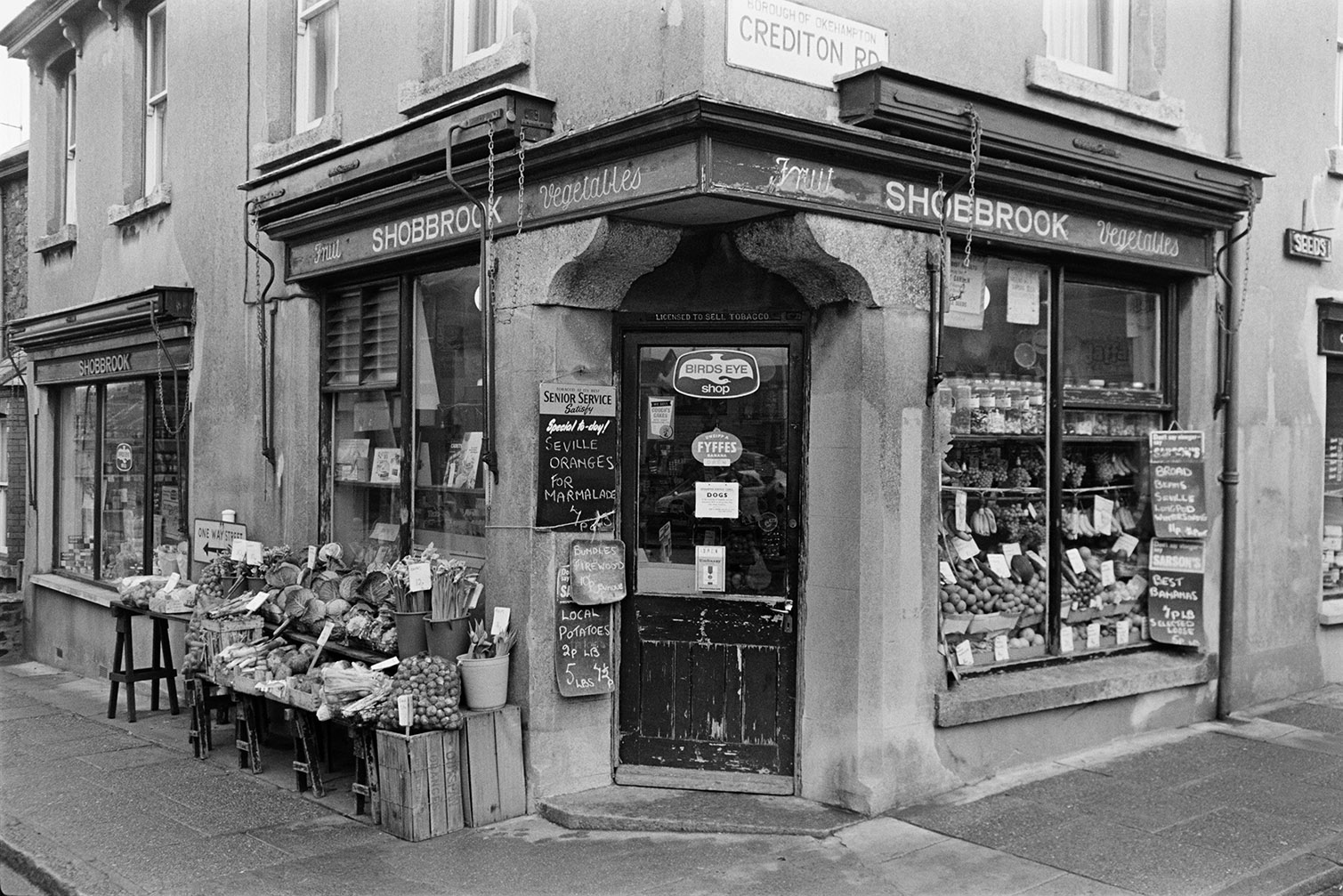 The Shop front window and display of Shobbrook fruit and vegetable corner shop on Crediton Road in Okehampton. Produce, including cabbages, daffodils and swedes, is on display outside the shop. More fruit and vegetables are displayed inside the shop window and two signs by the doorway are advertising Seville oranges and local potatoes.
