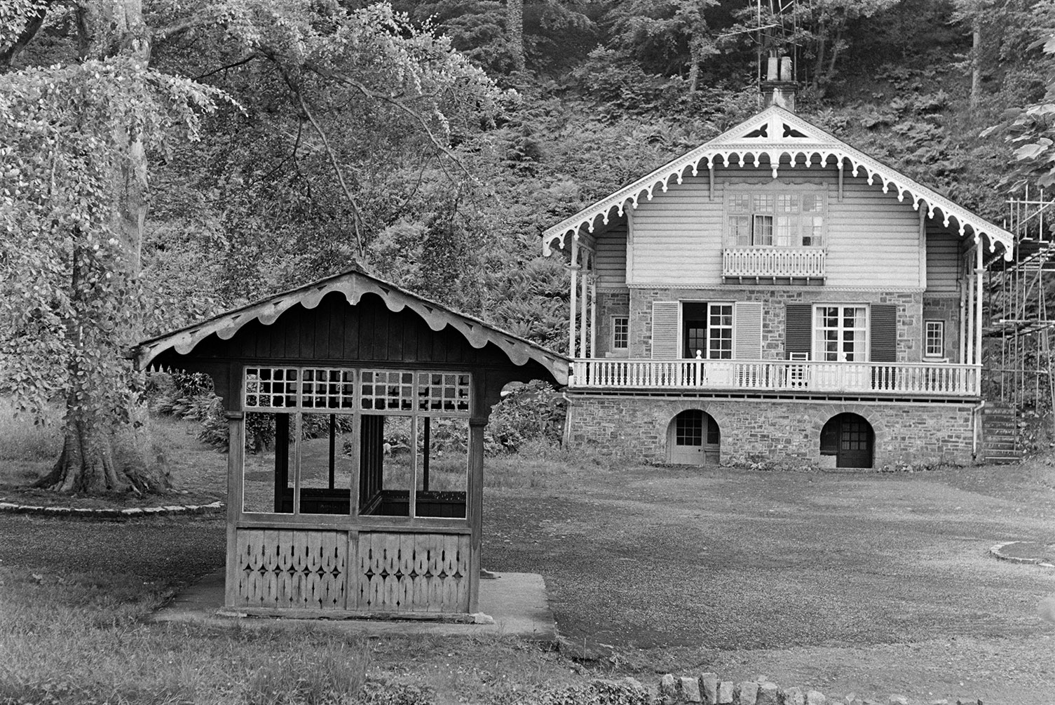 An alpine cottage at Simmons Park, Okehampton. A bench with a canopy is in the foreground and the cottage is surrounded by trees.