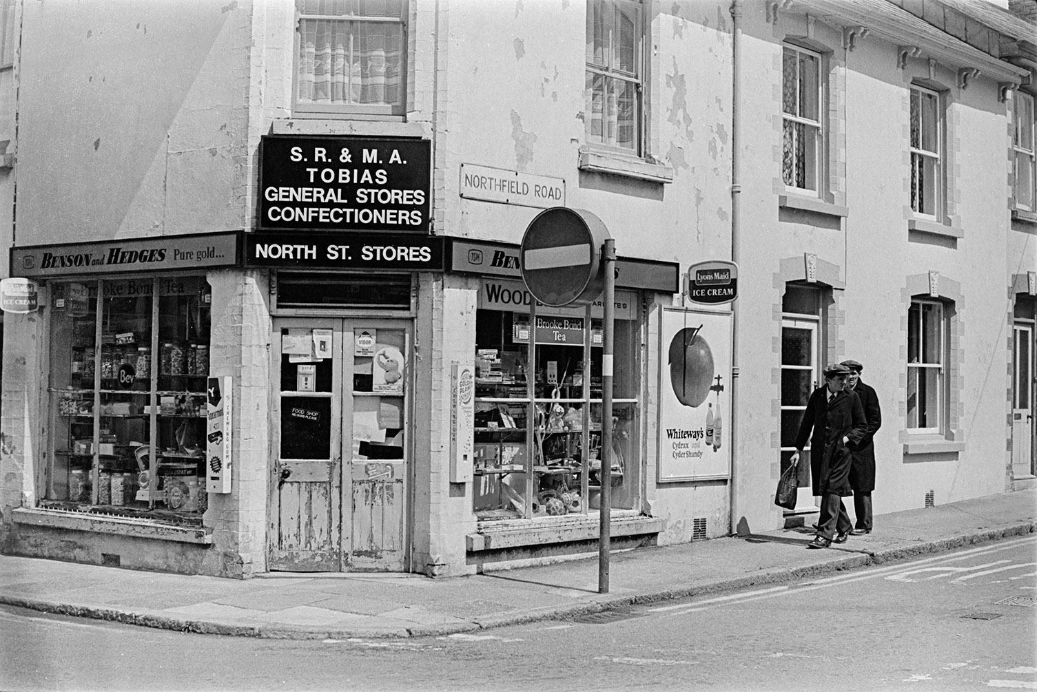 Two men walking towards North Street Stores on the corner shop of Northfield Road in Okehampton. The name above the shop is 'S R & M A Tobias'. Goods, including jars of sweets, are displayed in the shop front window.
