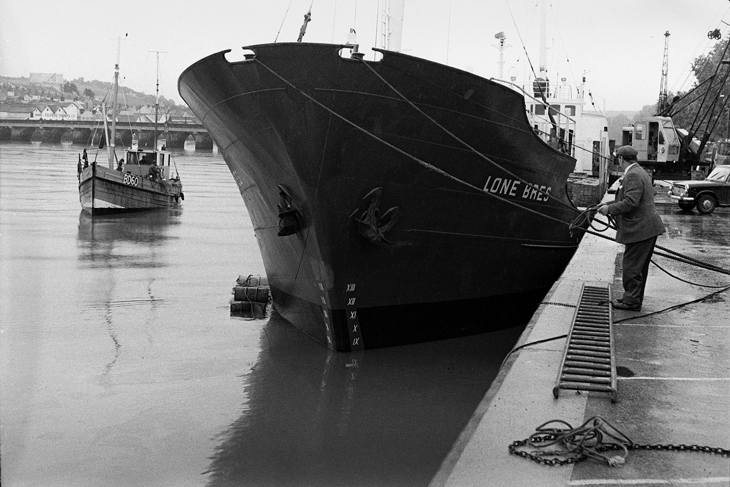 The ship 'Lone Bres' moored against the quayside at Bideford with a man stood by a ladder and holding ropes attached to the ship. A crane is on the quay ready to unload her cargo. A fishing boat can be seen on the River Torridge in the background, as well as Bideford Long Bridge, also known as Bideford Old Bridge.