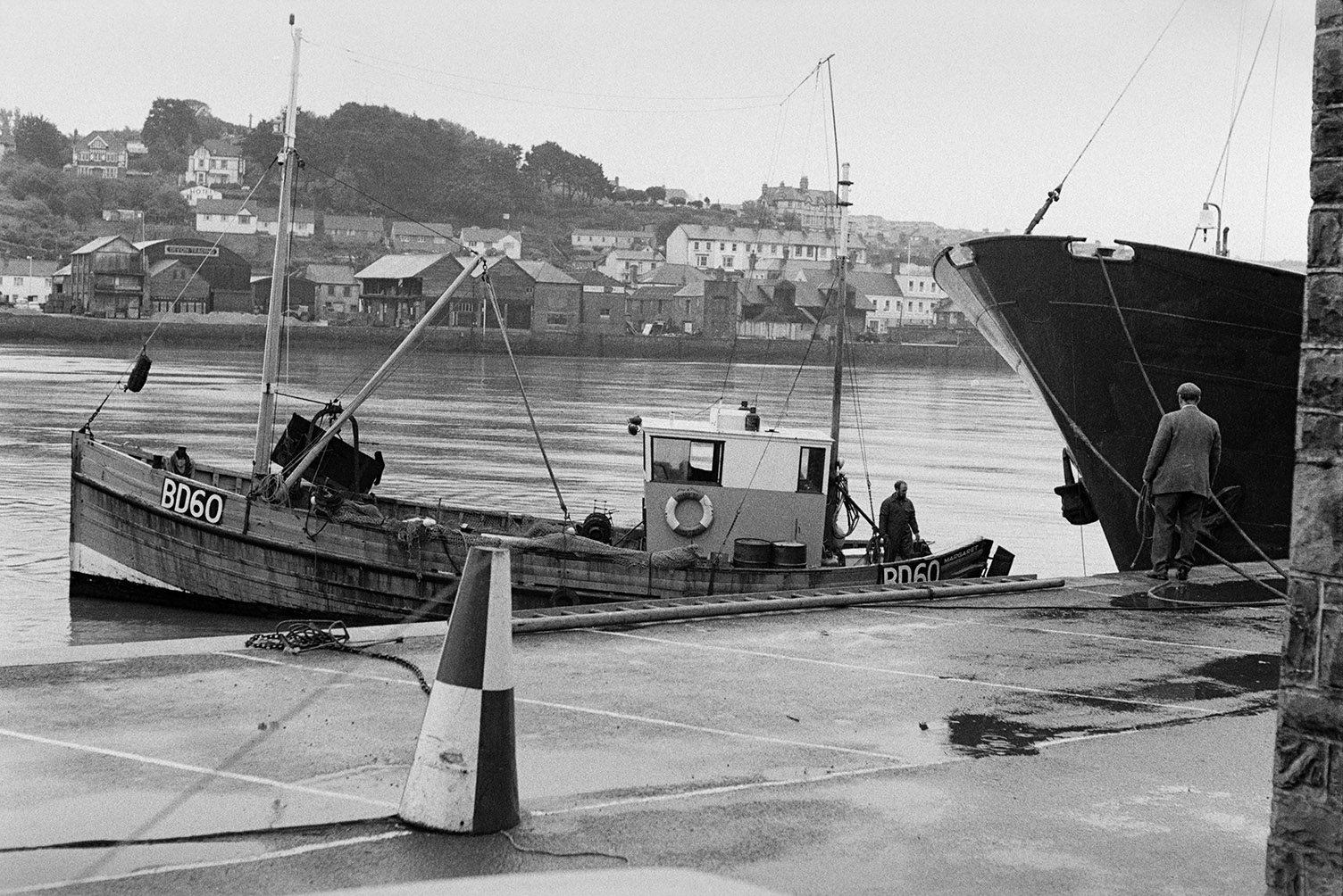 A fishing boat coming to moor up at the quayside at Bideford, next to the ship 'Lone Bres'. A man is waiting on the quayside with ropes to tie the boat up. Various building can be seen on the opposite side of the River Torridge.