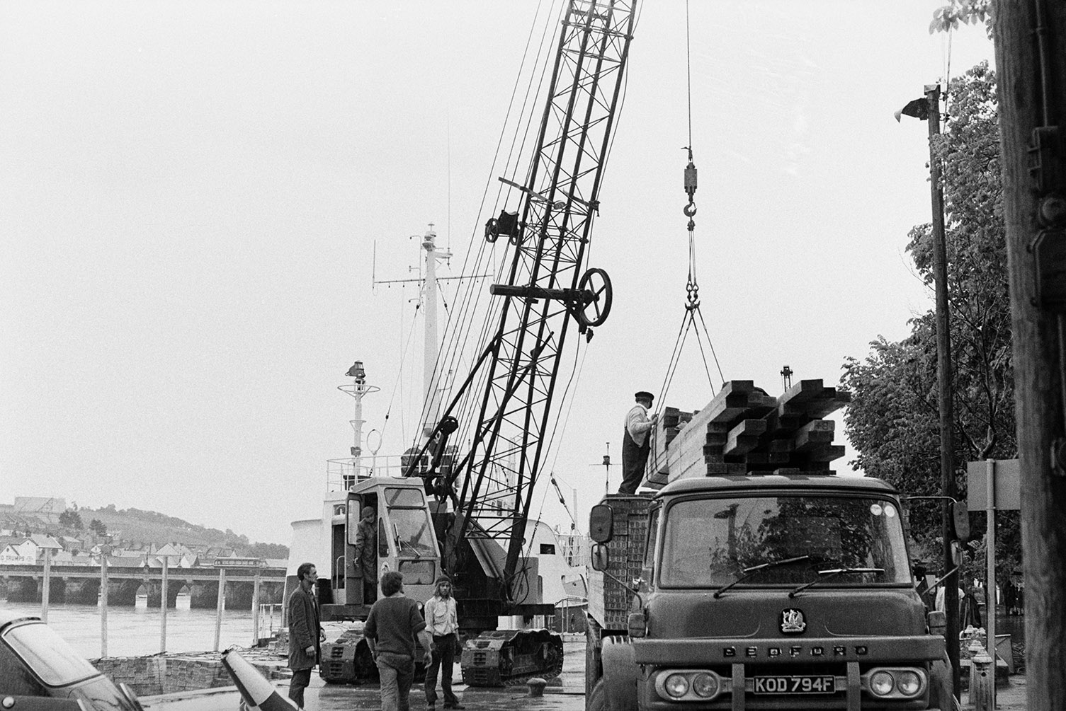 A crane loading planks of wood, taken from the ship 'Lone Bres' onto a lorry at Bideford Quay. Bideford Long Bridge, also known as Bideford Old Bridge can be seen in the background.