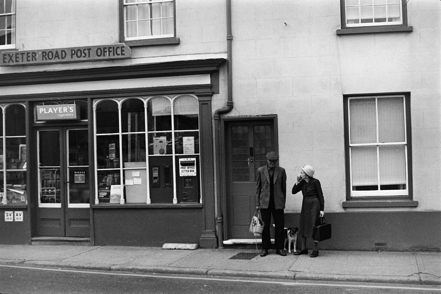 The shop front of the Exeter Road Post Office, in Crediton. A post box is set into the shop window and a man and woman are talking on the street outside with a dog.