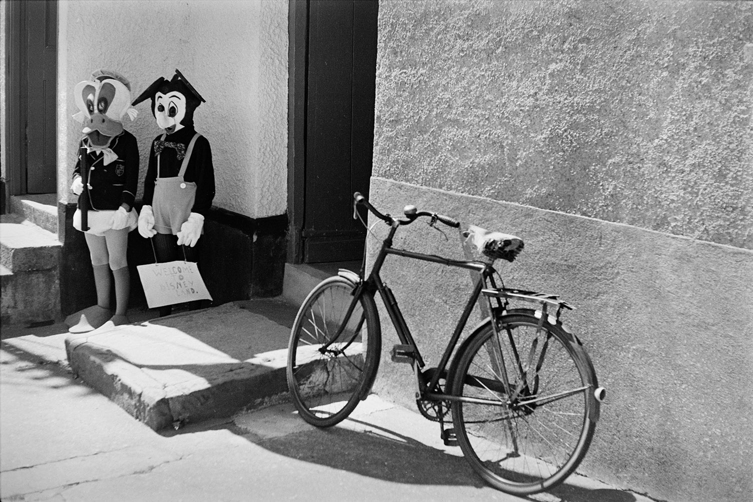Two children in fancy dress Disney characters Micky Mouse and Donald Duck at Winkleigh Fair. They are stood by a doorway with a bicycle propped up outside.