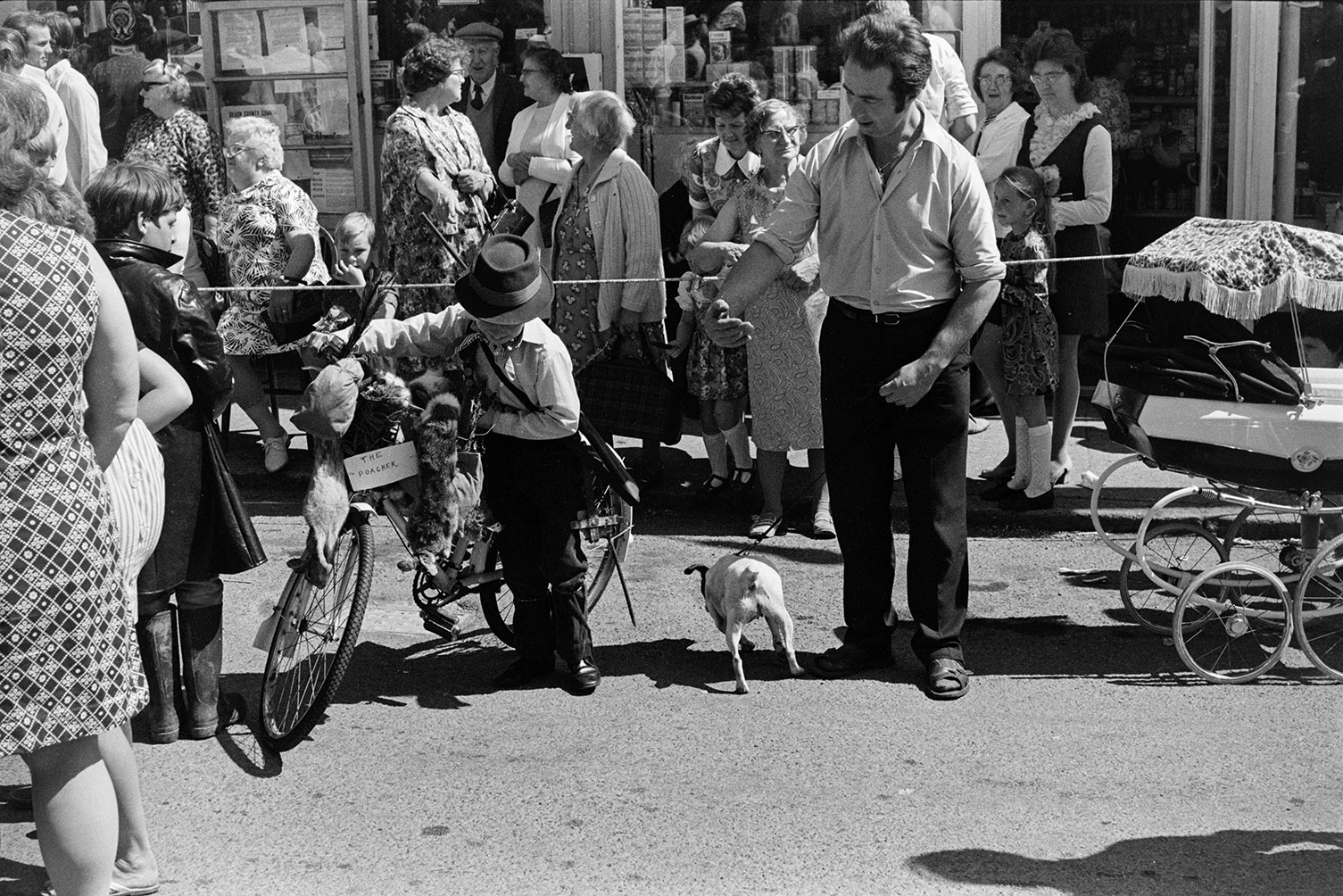 Spectators outside a shop front at Winkleigh Fair. A boy with a bicycle in a fancy dress costume titled 'The Poacher' is being guided by a man with a dog.