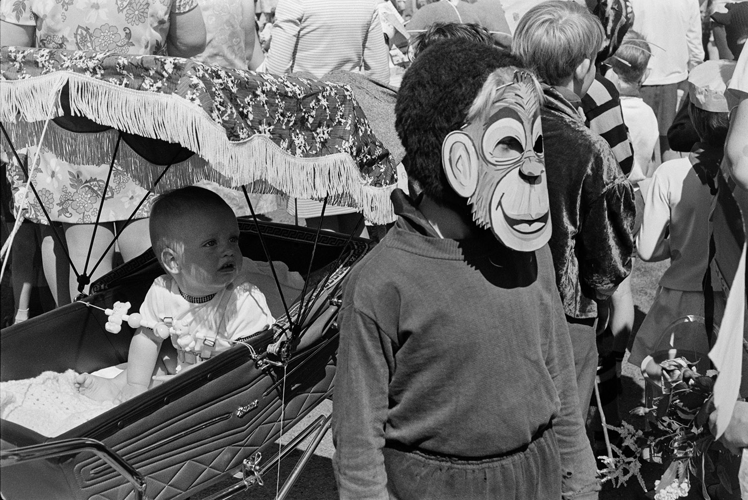 A child wearing a monkey mask stood in front of a pram with a baby in it at Winkleigh Fair.