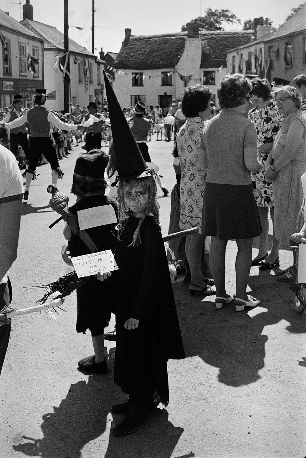A girl in fancy dress costume titled 'The Wicked Witch of Winkleigh' at Winkleigh Fair with other spectators. Another child behind her is dressed as Robinson Crusoe. The square is decorated with bunting.
