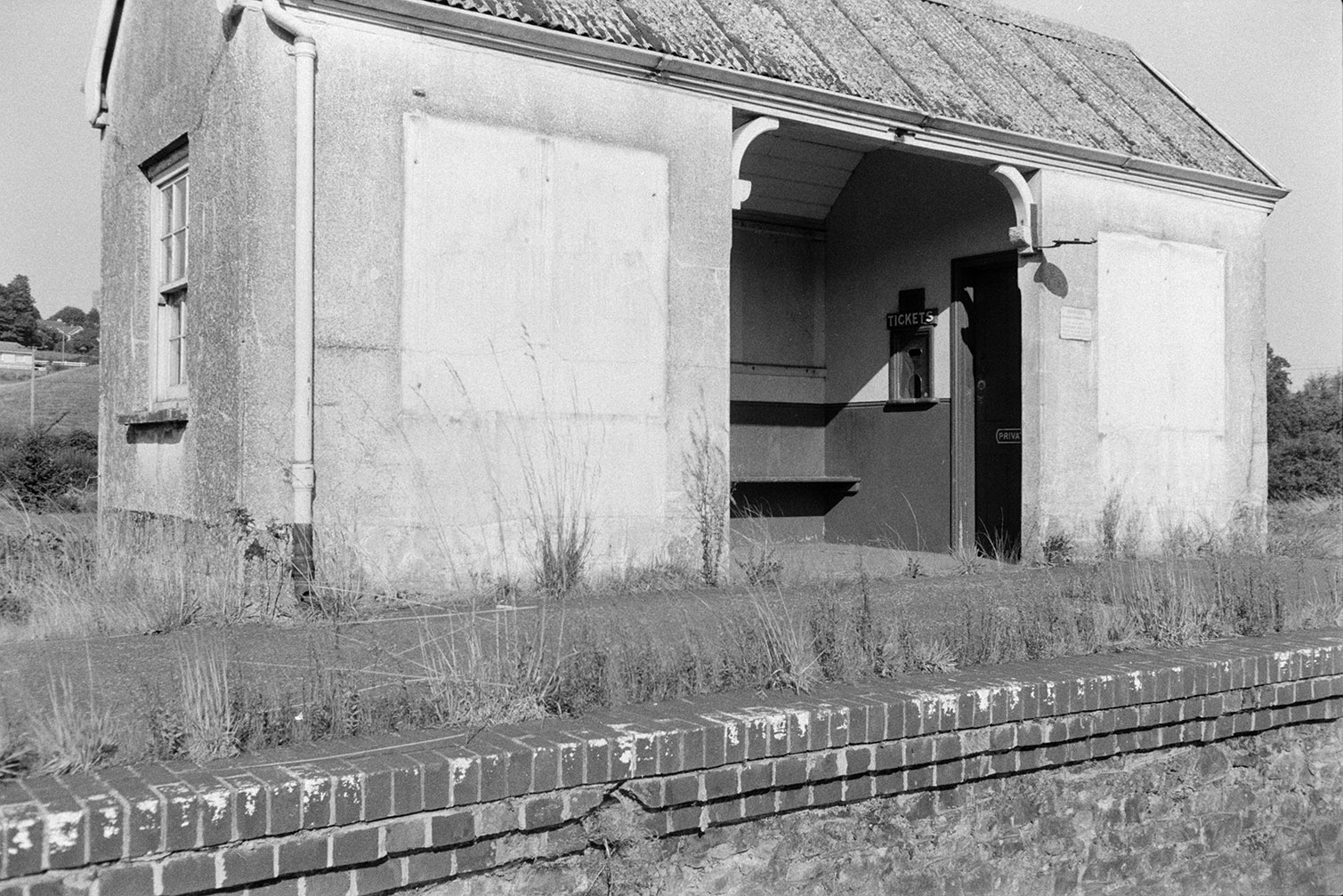 Railway platform, possibly at Lapford Railway Station. There is a shelter with a corrugated iron roof which has a ticket booth inside. The edge of the platform is overgrown with weeds.