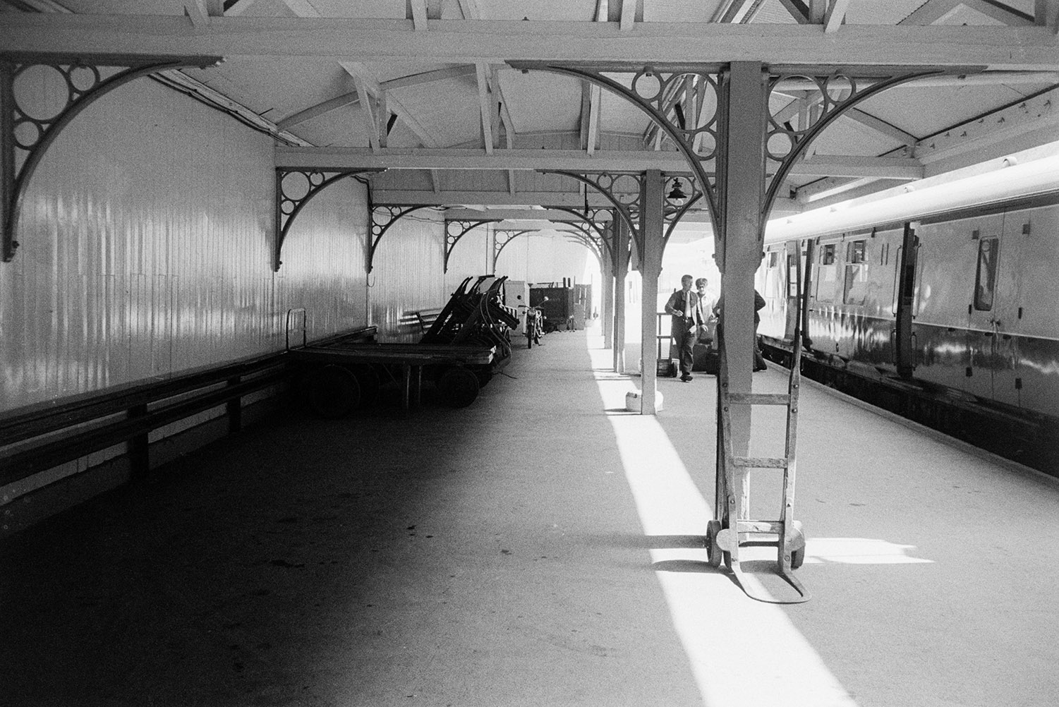 A train waiting at a platform at Barnstaple Railway Junction. Passengers are on the platform. Various other items, including two sack trucks and a bicycle can also be seen on the platform.