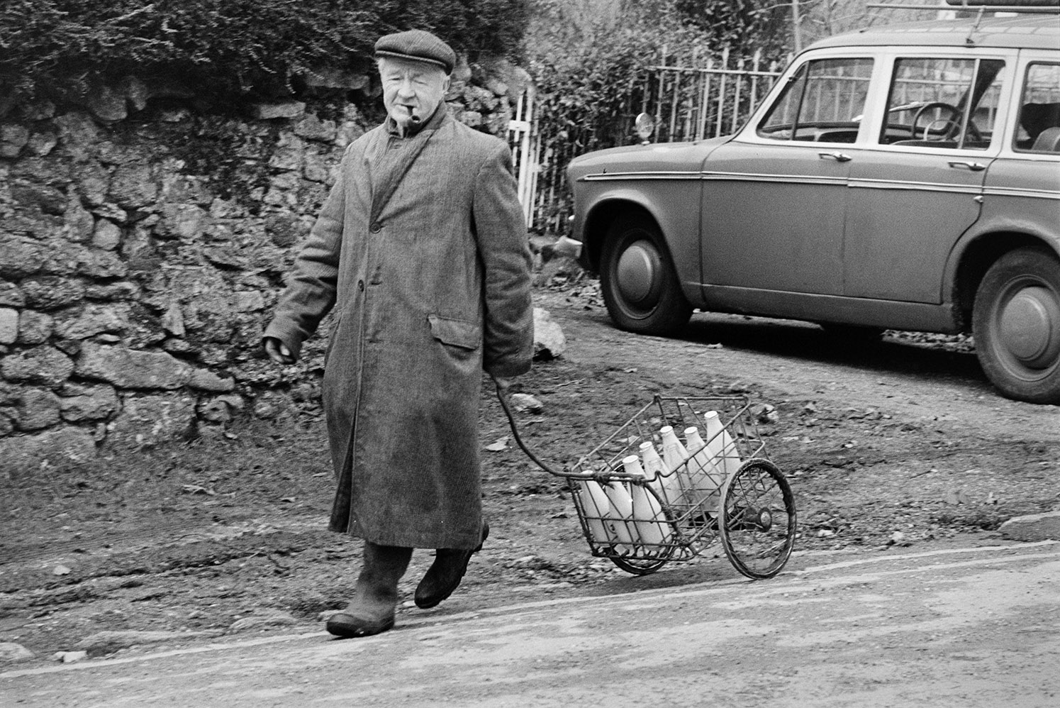 A man delivering milk on Dartmoor, using a small cart. He is walking past a parked car and stone wall, and is smoking a pipe.