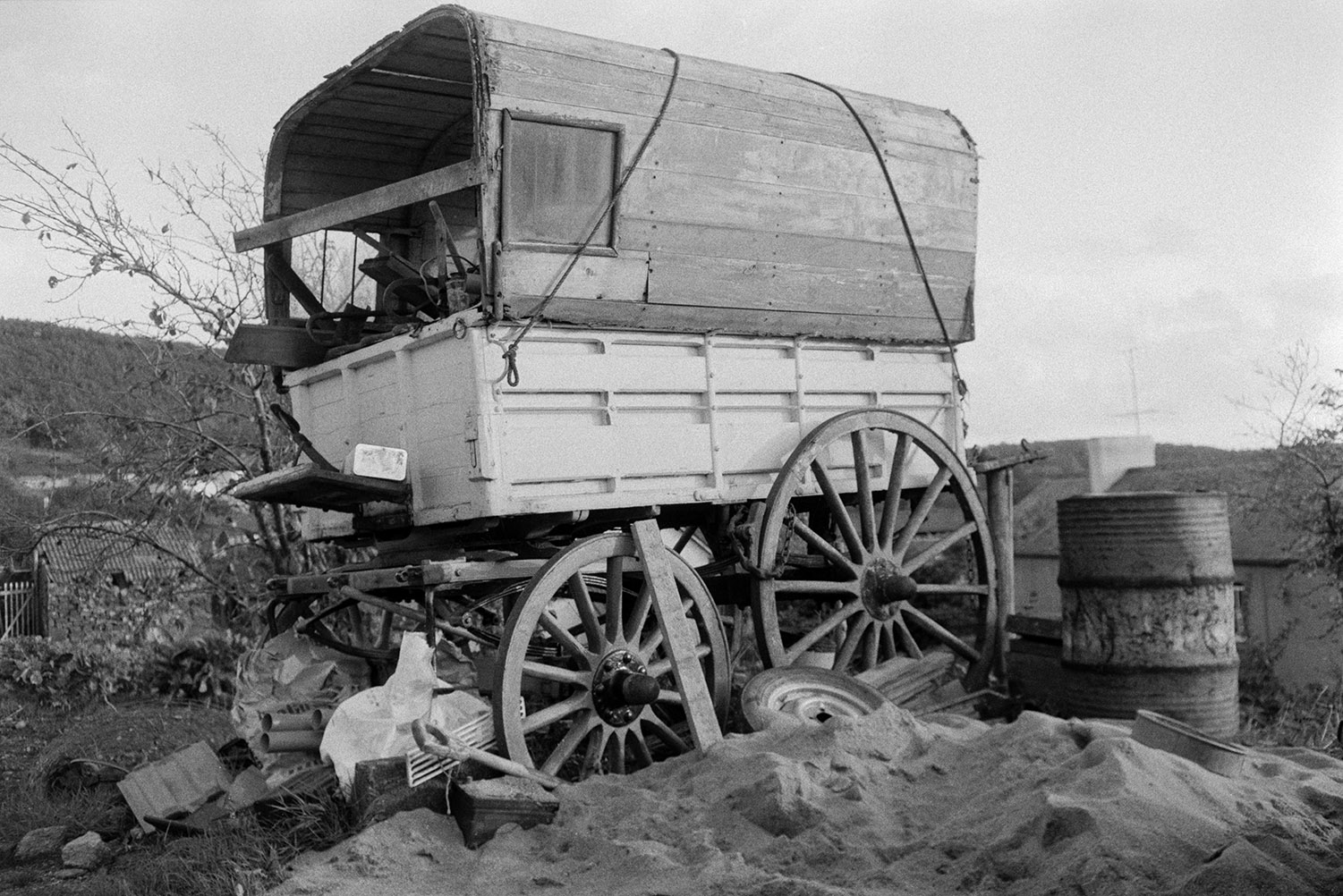 An old wagon with a wooden cover surrounded by an oil drum, sand and other items, at Weare Giffard. Its is possibly an old gypsy caravan.