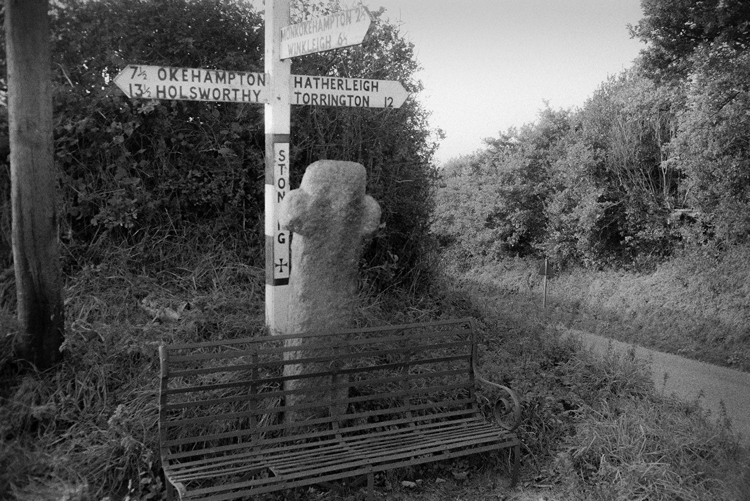 A signpost at Stoneleigh crossroads at Hatherleigh, behind a wayside cross and a metal bench.