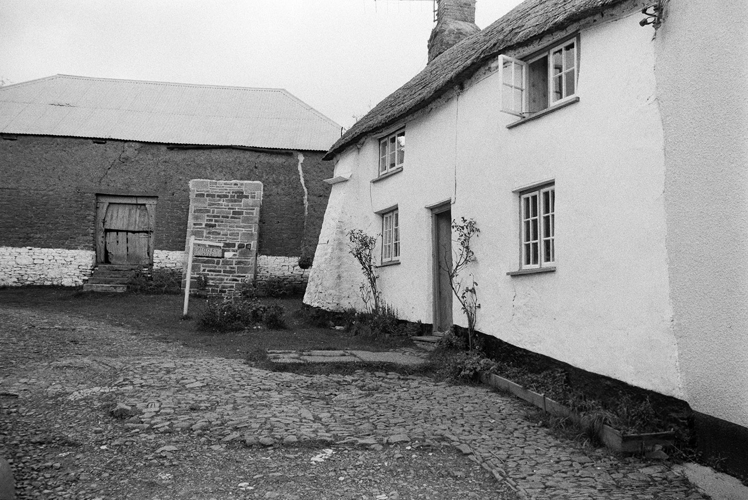 A thatched cottage next to a cob and stone barn with a corrugated iron roof, in Atherington. A sign outside the cottage reads 'Honey Sale' and the ground is cobbled.