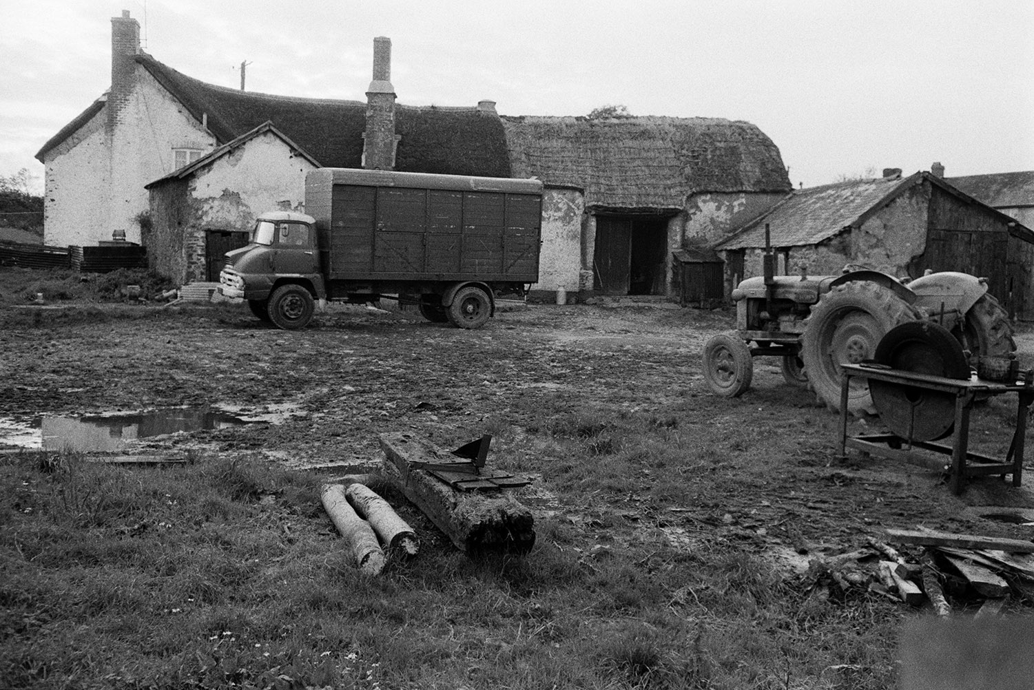 A farmyard with a tractor, circular saw, barns and a small lorry, in Atherington. The thatched farmhouse can be seen in the background.